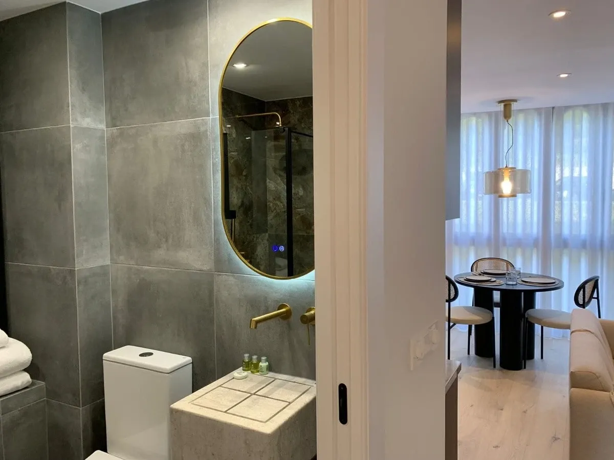 Baño con espejo ovalado iluminado y, al fondo, el salón del piso